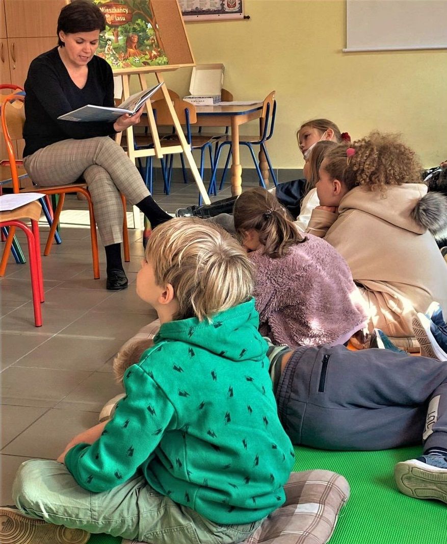Dzieci siedzą lub leżą na matach i słuchają opowieści czytanej przez kobietę.