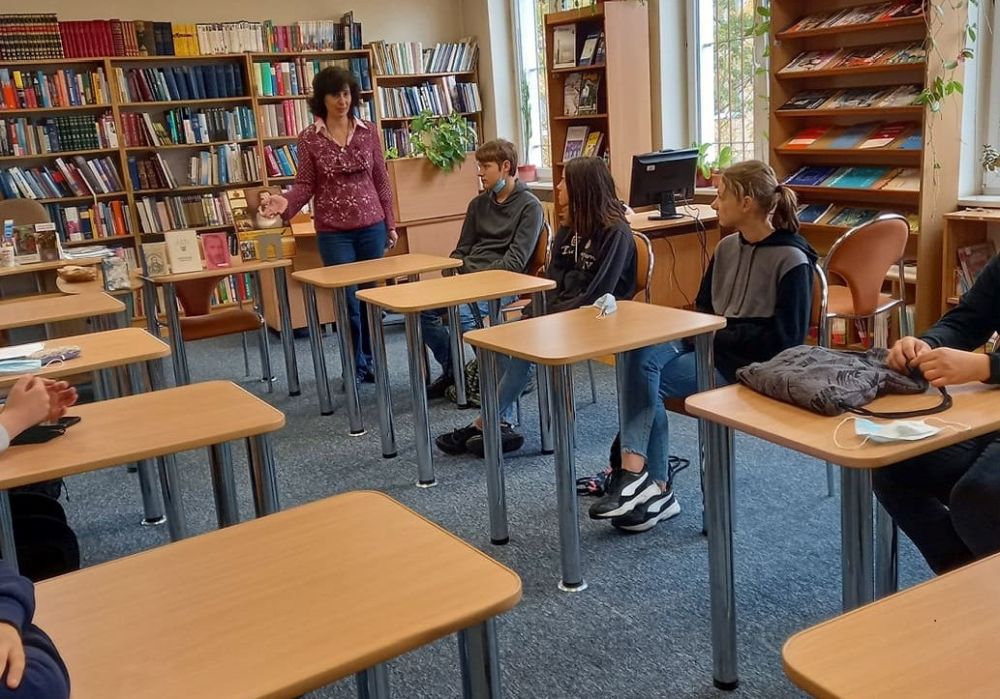 Bibliotekarka na tle ragałów z książkami w Czytelni prezentuje wybrane książki. Uczniowie siedza przy pojedynczych stolikach.
