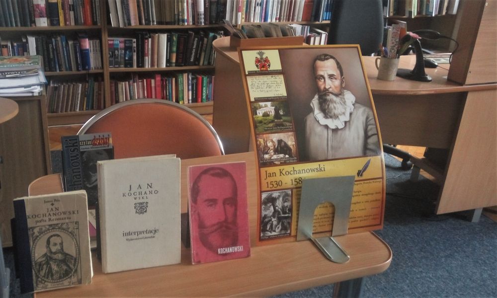 Książki i plakat o Janie Kochanowskim ustawione na stoliku. W tle regały z ksiązkami.