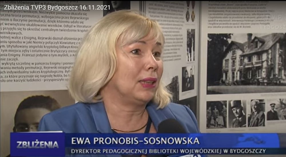 Dyrektor biblioteki Ewa Pronobis-Sosnowska mówi do mikrofonu TVP Bydgoszcz. W tle wystawa o Marianie Rejewskim.