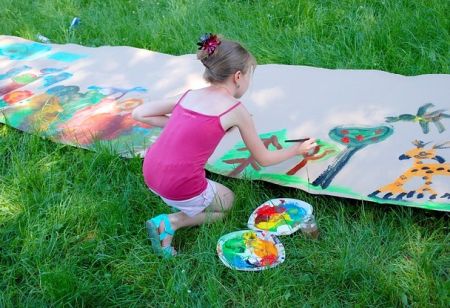 Dziewczynka maluje farbami na dużym arkuszu papieru rozłożonym na trawie.
