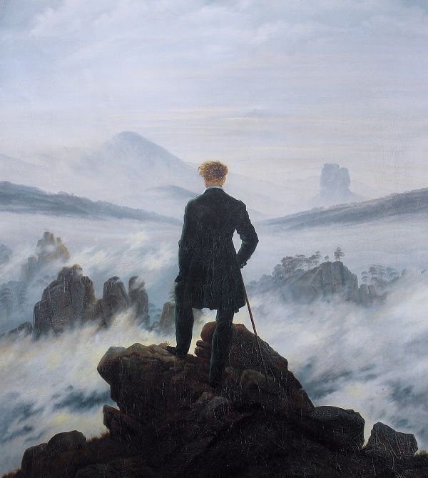 Mężczyzna w ciemnym surducie z laską stoi na szczycie skały. Przed nim zamglone szczyty i doliny.