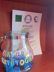 Szklany pojemnik z korkami i napisem "Lekturowa kryptogra". W tle dyplom za ukończenie gry.