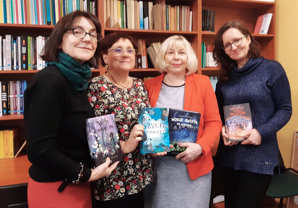 Cztery uśmiechnięte kobiety z książkami na tle bibliotecznych regałów.