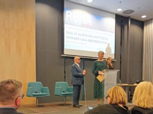 Dwie kobiety i niewidomy mężczyzna z białą laską przy mównicy i ekranie w sali konferencyjnej.