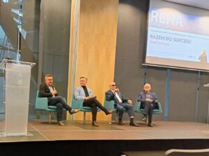 Czterech mężczyzn siedzi w fotelach przy mównicy i ekranie w sali konferencyjnej. Dwóch z nich trzyma mikrofony.