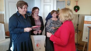 Kobieta w czerwonym żakiecie odbiera medal i upominki z rąk trzech kobiet.