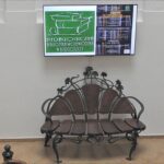 Stylowa ławka ze zdobieniami metalowymi. Nad nią ekran z logo biblioteki i programem seminarium.