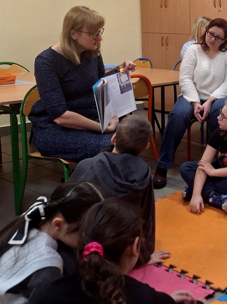 Kobieta prezentuje książkę dzieciom siedzącym na kolorowych matach.