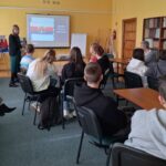 Młodzież siedzi przy stolikach i kieruje wzrok na kobietę stojącą przy ekranie z flagą Bydgoszczy.