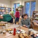 Kilka osób pry stoliku z ciasteczkami i napojami. W tle regały z książkami i czasopismami.