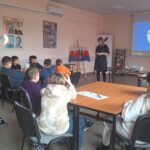 Dzieci przy stolikach słuchają kobiety, która stoi przy ekranie z napisem "O czym szumi Brda?"