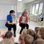 W przedszkolu dzieci zwrócone są w kierunku dwóch kobiet. Jedna pokazuje książkę.