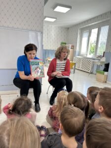 W przedszkolu dzieci zwrócone są w kierunku dwóch kobiet. Jedna pokazuje książkę.