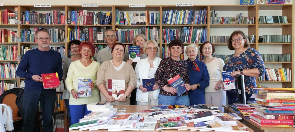 Seniorzy pozują z książkami na tle regałów bibliotecznych.