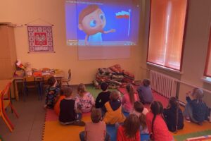 Małe dzieci siedzą na kolorowych matach i oglądają film o barwach narodowych.