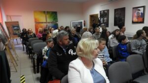 Sala z obrazami na ścianach. W środku grupa ludzi na krzesłach zwrócona w jedną stronę.