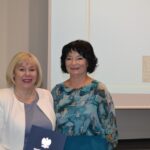 Kobieta w białej marynarce i kobieta w turkusowej sukience uśmiechają się.