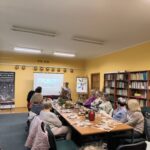Sala wykładowa. Regały z książkami. Prezentacja w tle. W centrum stół z poczęstunkiem, wokół na krzesłach ludzie.