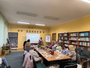 Sala wykładowa. Regały z książkami. Prezentacja w tle. W centrum stół z poczęstunkiem, wokół na krzesłach ludzie.