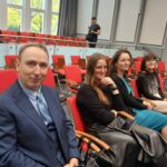 4 osoby, siedzą na krzesłach i się uśmiechają.