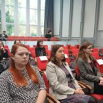 Rudowłosa kobieta w koszuli w kropki, kobieta w jasnym garniturze, kobieta w brązowym komplecie. W tle kilka innych osób.