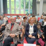 W centrum mężczyzna w jasnej marynarce i kobieta w białej koszuli. W tle inne osoby.