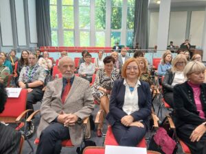 W centrum mężczyzna w jasnej marynarce i kobieta w białej koszuli. W tle inne osoby.