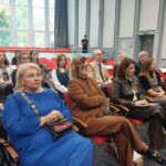 Kolejno: kobieta w niebieskiej sukni, kobieta w brązowym garniturze, kobieta w czarnym garniturze. W tle inne osoby.