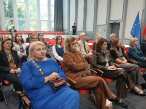 Kolejno: kobieta w niebieskiej sukni, kobieta w brązowym garniturze, kobieta w czarnym garniturze. W tle inne osoby.