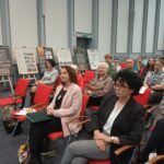 Dwie kobiety w pierwszym rzędzie, w tle kilka innych postaci oraz wystawa.