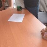 Blat stołu, z dwoma małymi kartonikami oraz nazwą gry. Wokół kilka osób.