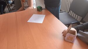 Blat stołu, z dwoma małymi kartonikami oraz nazwą gry. Wokół kilka osób.