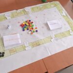 Papierowa plansza z polami gry, dwiema stertami kart z zadaniami i guzikami