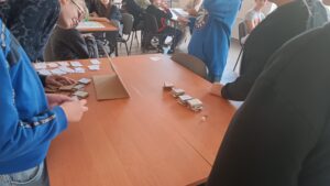 Stolik, na blacie kartonowe elementy gry, wokół młodzież.