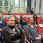 Kobieta ubrana w czarną sukienkę i kobieta w koszuli we wzory siedzą na sali. W tle kilka innych osób.