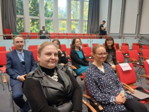 Kobieta ubrana w czarną sukienkę i kobieta w koszuli we wzory siedzą na sali. W tle kilka innych osób.