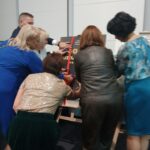 Siedem osób stoi lub pochyla się nad sztalugą - tyłem do obiektywu.