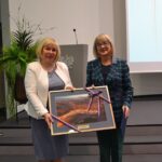 Kobieta w białej marynarce i kobieta w niebieskim żakiecie trzymają obraz.