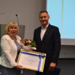 Kobieta w białej marynarce i mężczyzna w ciemnym garniturze trzymają ramkę owiniętą szarfą.