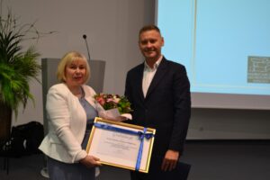 Kobieta w białej marynarce i mężczyzna w ciemnym garniturze trzymają ramkę owiniętą szarfą.