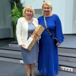 Kobieta w białej marynarce i kobieta w niebieskiej sukni z kartonową paczką.