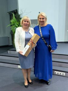 Kobieta w białej marynarce i kobieta w niebieskiej sukni z kartonową paczką.