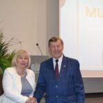 Kobieta w białej marynarce i mężczyzna w granatowym garniturze w trakcie uścisku dłoni.