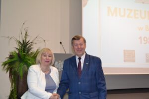 Kobieta w białej marynarce i mężczyzna w granatowym garniturze w trakcie uścisku dłoni.