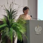 Kobieta w złotej bluzce za katedrą z orłem.