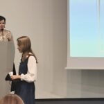 Kobieta w czarnej sukience wręcza nagrodę dziewczynce w białej koszuli i sukience. Za nimi kobieta na podium.