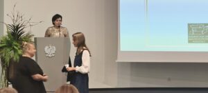 Kobieta w czarnej sukience wręcza nagrodę dziewczynce w białej koszuli i sukience. Za nimi kobieta na podium.