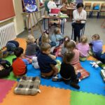 Grupa siedzących dzieci słucha czarnowłosej siedzącej na krześle kobiety.