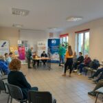 Sala pełna ludzi siedzących na krzesłach, kobieta w żółtej koszuli stoi w centrum.